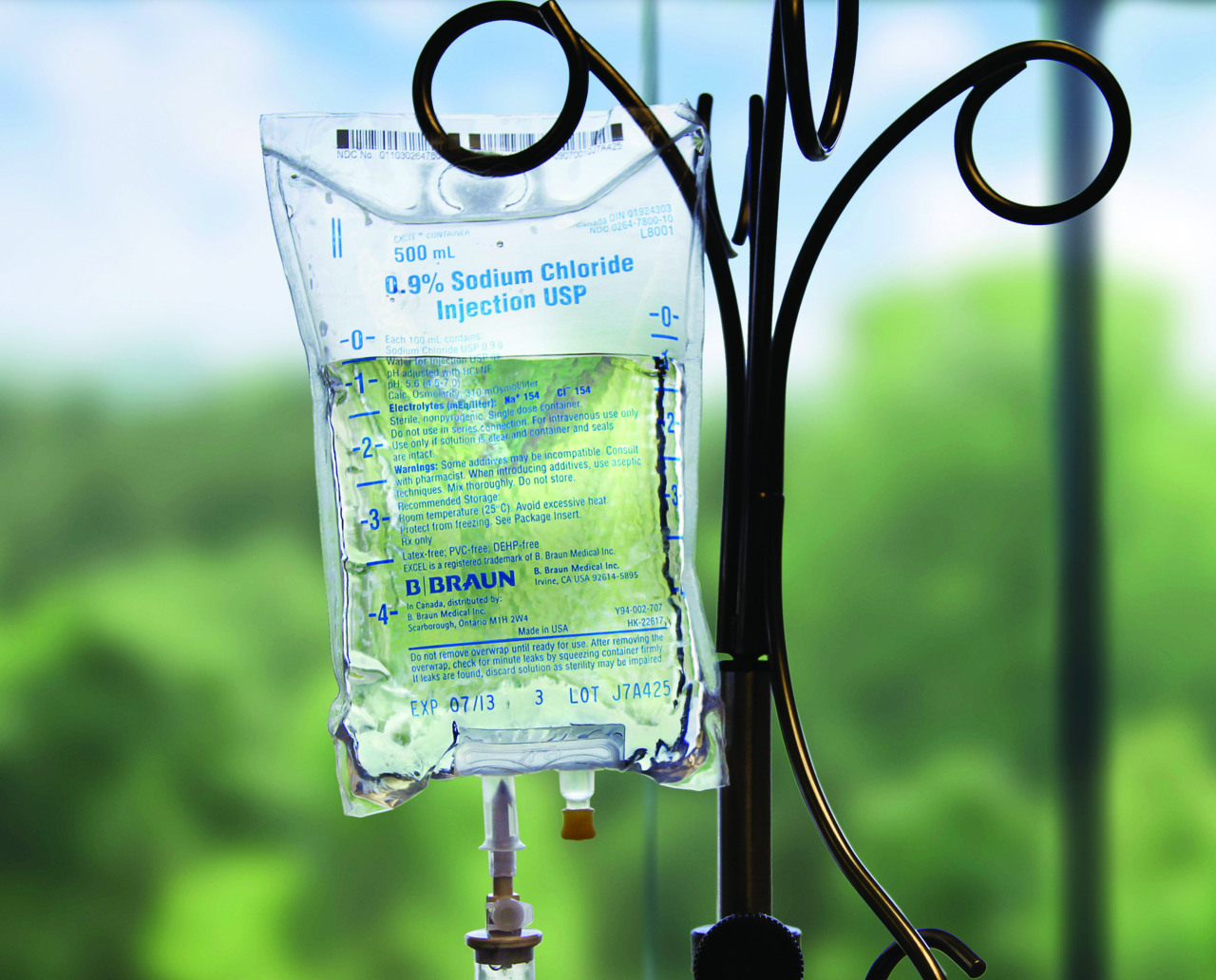 An IV container with 0.9% Sodium Chloride Injection USP being held up by a gloved hand in front of a window looking outside.