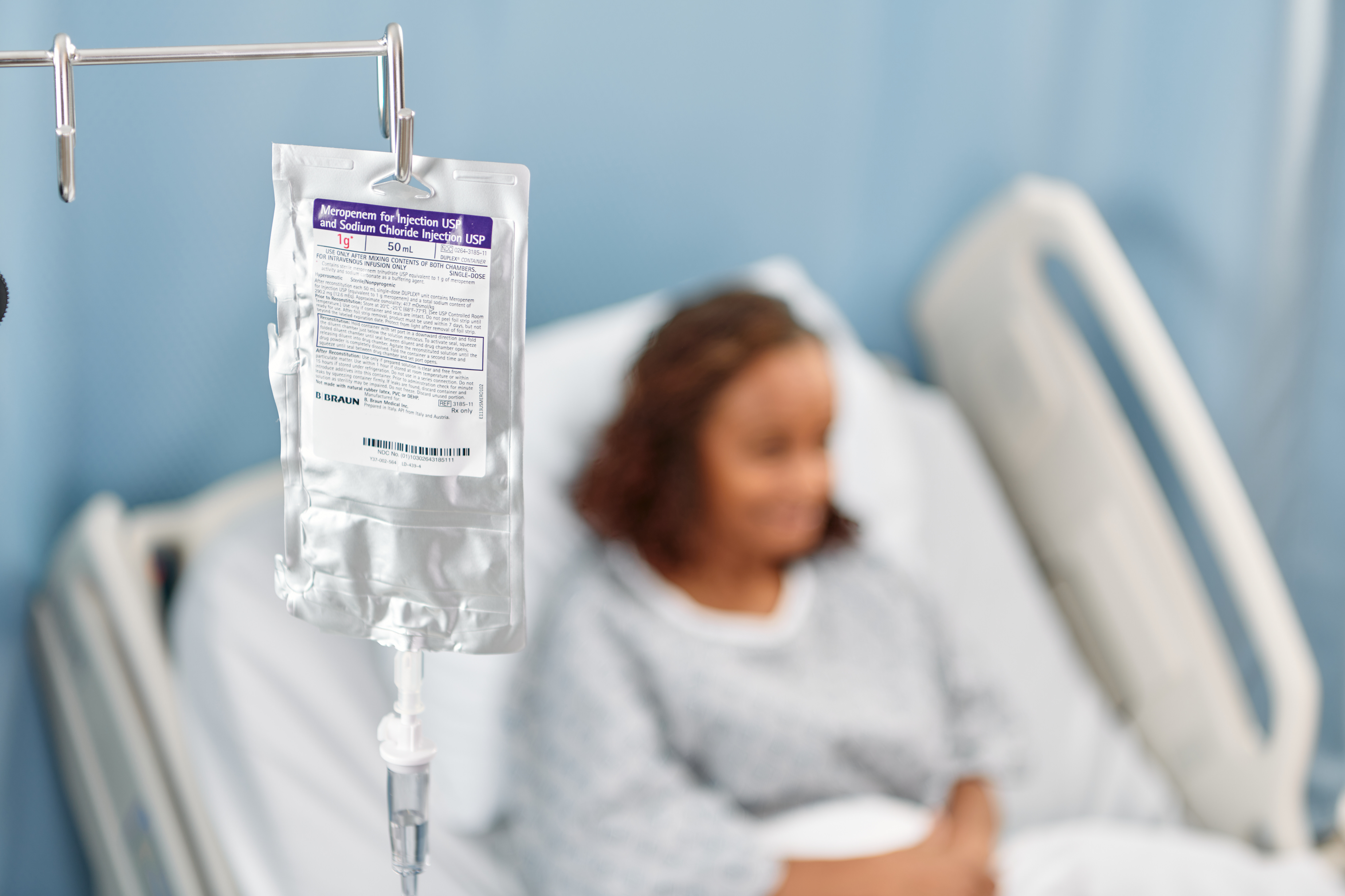 Patient sitting on a hospital bed in the background with a DUPLEX meropenem container handing in the foreground
