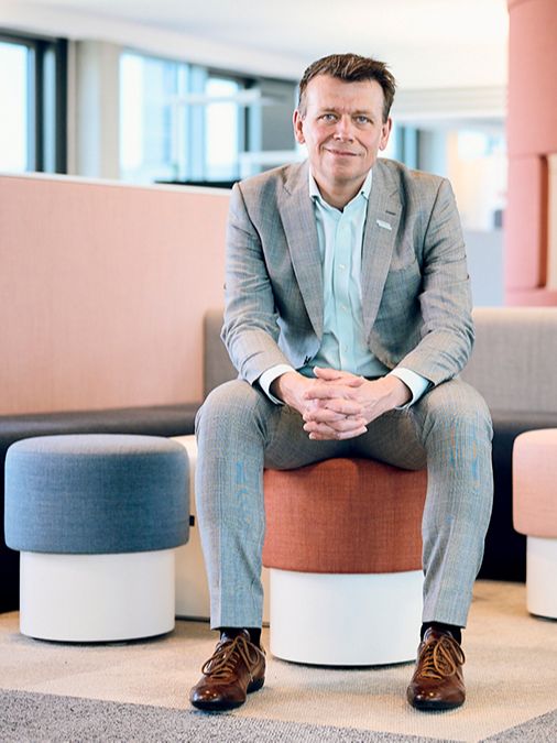Dr. Michael Lauk sitting on a red fabric stool in a modern office, smiling and with folded hands. 