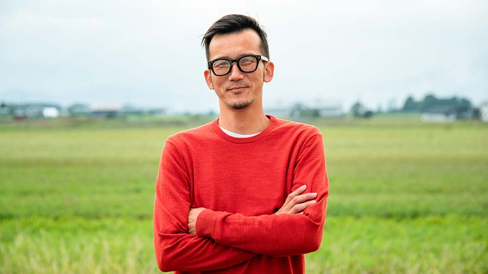 Une personne portant des lunettes se tient debout avec confiance et les bras croisés dans un vaste champ.